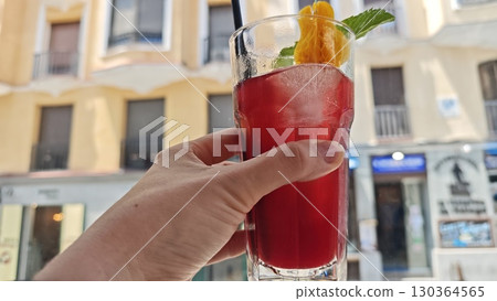A hand holding a glass of red sangria with ice, orange slice and mint garnish against a sunny street background 130364565