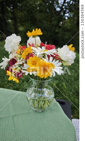 Mixed bouquet of summer flowers in a clear glass vase placed on a green tablecloth outdoors 130364569