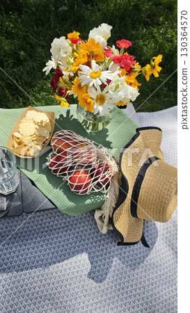 Colorful summer picnic setup with fresh flowers, peaches in a mesh bag, crackers and a straw hat on a blanket 130364570