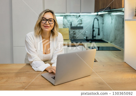 Freelance woman working on laptop in modern kitchen. Blonde adult in glasses and bathrobe focused on screen, remote work lifestyle, morning routine, cozy home office setup. 130365092