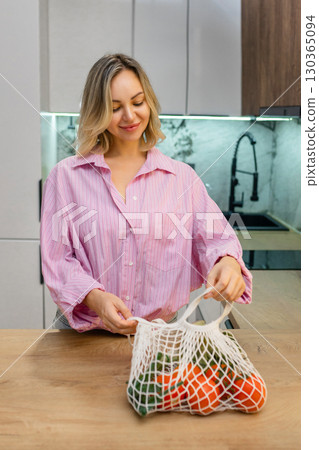 Smiling woman in pink shirt leaning on kitchen counter next to mesh bag with fresh vegetables. Healthy lifestyle and eco-friendly food shopping at home. Smiling woman in pink shirt leaning on kitchen counter next to mesh bag with fresh vegetables. Healthy lifestyle and eco-friendly food shopping at home. 130365094