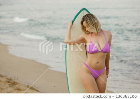 Attractive young woman with prominent cleavage in purple bikini standing with surfboard on sandy beach by the sea during sunset. Summer vacation and surfing lifestyle Attractive young woman with prominent cleavage in purple bikini standing with surfboard on sandy beach by the sea during sunset. Summer vacation and surfing lifestyle 130365100