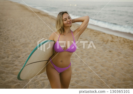 Attractive young woman in a purple bikini posing with a surfboard on the beach, enjoying summer by the sea, confident and relaxed, ocean waves in the background. Attractive young woman in a purple bikini posing with a surfboard on the beach, enjoying summer by the sea, confident and relaxed, ocean waves in the background. 130365101