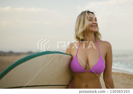 Attractive young woman in a purple bikini posing with a surfboard on the beach, enjoying summer by the sea, confident and relaxed, ocean waves in the background. 130365103