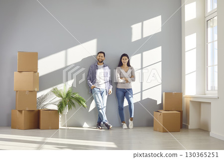 Happy family couple in casual clothes are standing together near wall in rays of sunlight among cardboard boxes. Married smiling couple buys or rents new house. Real estate, relocation, own housing. 130365251