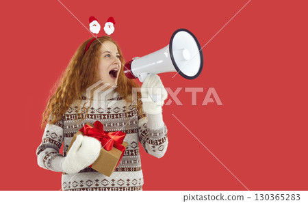 Happy redhead girl holding Christmas gift present in hands shouting loudspeaker on red background. 130365283