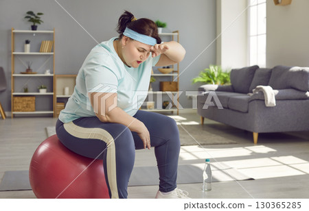 Fat woman, tired after workout, sitting on exercise ball and wiping sweat off her forehead 130365285