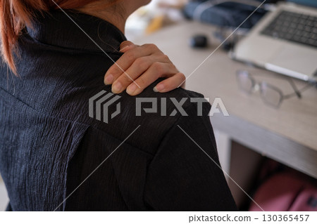 Hands rubbing aching shoulders in office setting with laptop and work equipment nearby 130365457