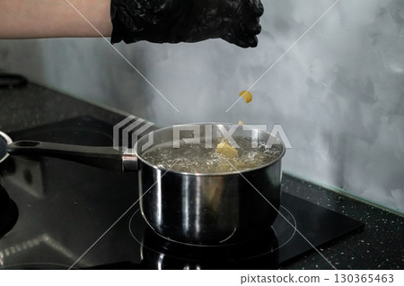 Gloved hand dropping pasta into a pot of boiling water on an induction hob, symbolizing home cooking and meal preparation in a modern kitchen setting Gloved hand dropping pasta into a pot of boiling water on an induction hob, symbolizing home cooking and meal preparation in a modern kitchen setting 130365463