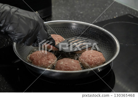 Person in black glove frying raw cutlets with a spatula in a hot pan on an induction stove for home cooking, healthy meal preparation 130365468