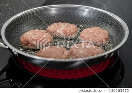 Fresh raw meat cutlets sizzling in hot oil within a patterned frying pan on an induction cooktop, symbolizing delicious homemade meal preparation and cooking 130365475