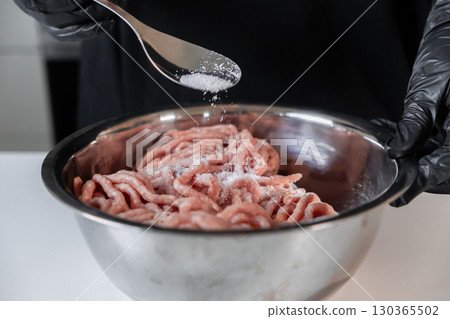Professional chef in black protective gloves carefully seasons raw minced meat with salt in a stainless steel bowl, preparing savory cutlets for a home culinary experience 130365502