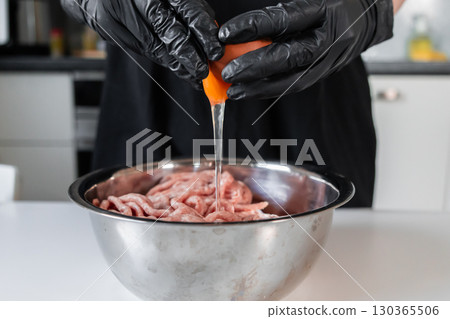 Chef in black gloves cracking a fresh egg into a bowl of raw minced meat, preparing ingredients for making delicious homemade cutlets 130365506