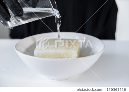Gloved hand pouring water over a block of bread soaking in a white bowl, essential for preparing a traditional culinary recipe Gloved hand pouring water over a block of bread soaking in a white bowl, essential for preparing a traditional culinary recipe 130365516