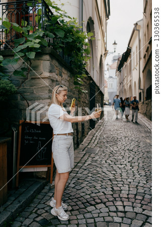 Young blonde woman taking picture of glass of white wine with her smartphone in narrow cobbled street of old town. Blonde woman photographing white wine glass near historic cobblestone alley Young blonde woman taking picture of glass of white wine with her smartphone in narrow cobbled street of old town. Blonde woman photographing white wine glass near historic cobblestone alley 130365538