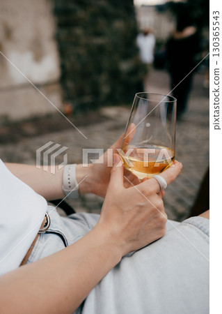 Woman sitting outdoors in casual attire, savoring glass of chilled white wine while enjoying peaceful moment of relaxation amidst vibrant urban backdrop. Woman relaxing outdoors, holding white wine Woman sitting outdoors in casual attire, savoring glass of chilled white wine while enjoying peaceful moment of relaxation amidst vibrant urban backdrop. Woman relaxing outdoors, holding white wine 130365543