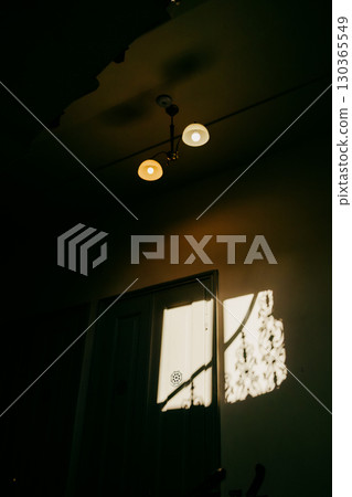 Sunlight casting decorative shadows on wall in dark room with ceiling lamp illuminating scene, creating mysterious and evocative atmosphere. Sunlight filtering through shadowy room, casting patterns Sunlight casting decorative shadows on wall in dark room with ceiling lamp illuminating scene, creating mysterious and evocative atmosphere. Sunlight filtering through shadowy room, casting patterns 130365549