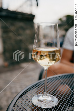 Glass of chilled white wine sits on metal table, reflecting surrounding outdoor setting, creating relaxed and inviting atmosphere. Chilled white wine glass resting on metallic surface. Wine tasting Glass of chilled white wine sits on metal table, reflecting surrounding outdoor setting, creating relaxed and inviting atmosphere. Chilled white wine glass resting on metallic surface. Wine tasting 130365554