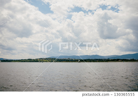 Calm lake water reflecting cloudy sky, showcasing distant mountains and charming lakeside village. Sailboats gracefully navigating serene landscape on warm summer day. Tranquil alpine lake Calm lake water reflecting cloudy sky, showcasing distant mountains and charming lakeside village. Sailboats gracefully navigating serene landscape on warm summer day. Tranquil alpine lake 130365555