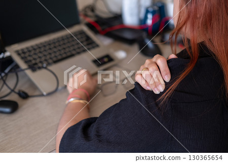Woman massaging sore shoulders at desk with laptop and office tools highlighting work fatigue Woman massaging sore shoulders at desk with laptop and office tools highlighting work fatigue 130365654