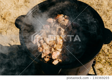 Pieces of meat frying in oil in a large cast iron cauldron with steam. Cooking outdoor in a big pot for holiday. 130369203