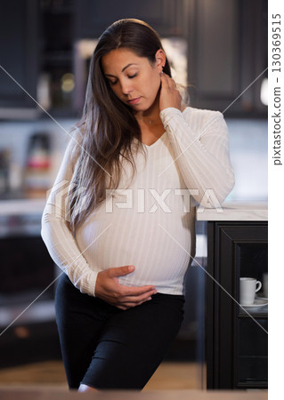 Pregnant woman in a white sweater standing thoughtfully in a kitchen 130369515