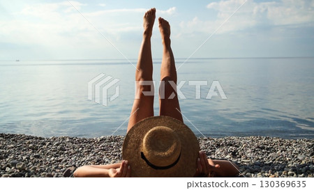 Beach Sunbathing Woman: Woman relaxes on pebble beach, legs up, sunny day by the sea, enjoying vacation. 130369635