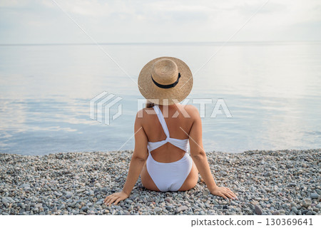 A woman wearing a white bikini and a straw hat is sitting on a beach. The beach is rocky and the sky is cloudy. A woman wearing a white bikini and a straw hat is sitting on a beach. The beach is rocky and the sky is cloudy. 130369641