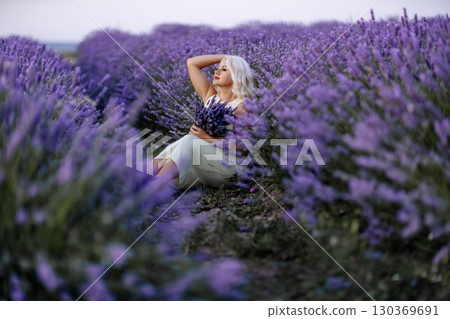 Blonde woman poses in lavender field at sunset. Happy woman in white dress holds lavender bouquet. Aromatherapy concept, lavender oil, photo session in lavender Blonde woman poses in lavender field at sunset. Happy woman in white dress holds lavender bouquet. Aromatherapy concept, lavender oil, photo session in lavender 130369691