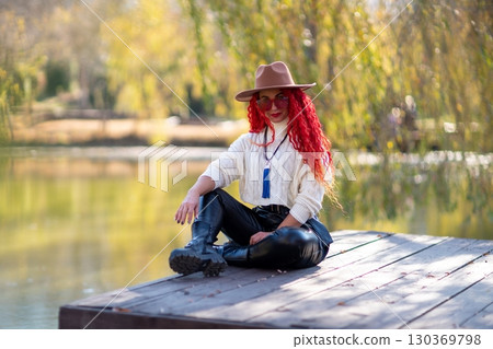 Autumn lake woman. In autumn, she sits by the pond on a wooden pier and admires nature with red hair and a hat. Tourism concept, weekend outside the city. 130369798