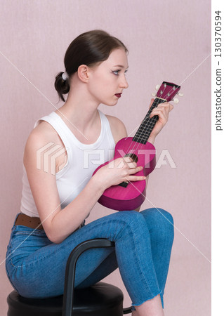 Serious young female musician playing ukulele, sitting on bar stool and looking away while plucking musical instrument. Profile view slender woman in casual clothing - white vest, blue skinny jeans Serious young female musician playing ukulele, sitting on bar stool and looking away while plucking musical instrument. Profile view slender woman in casual clothing - white vest, blue skinny jeans 130370594