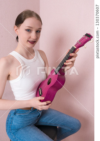 Young woman musician in positive light, holding ukulele. Her face conveys joy and she is looking at camera with sincere smile. Her positive facial expression conveys sense of cheer and friendliness 130370607