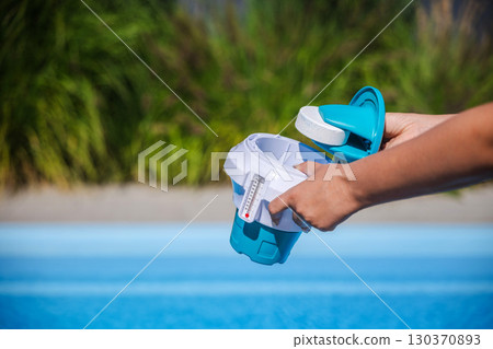 Hands placing chlorine tablet into pool dispenser with thermometer for clean and safe water maintenance Hands placing chlorine tablet into pool dispenser with thermometer for clean and safe water maintenance 130370893