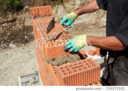 Skilled bricklayer constructs a sturdy wall using red bricks and mortar under bright sunlight in a construction site Skilled bricklayer constructs a sturdy wall using red bricks and mortar under bright sunlight in a construction site 130373101