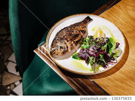 Grilled fish served with fresh greens and lemon slice in cozy dining setting Grilled fish served with fresh greens and lemon slice in cozy dining setting 130375655