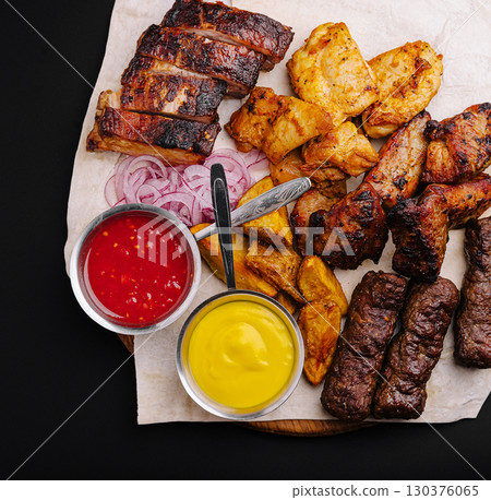 Grilled meat platter featuring ribs, chicken, and sausages with sauces 130376065