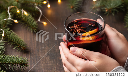Christmas mulled wine with orange slices and spices in hands on wooden table with fir branches and lights. Cozy winter holiday drink. 130381122