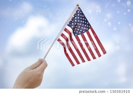 Human hand holding an American flag with a blue sky background. 4th of July concept. Close up on USA flag in a hand of a person. Independence Day or traveling in America concept. 130381910