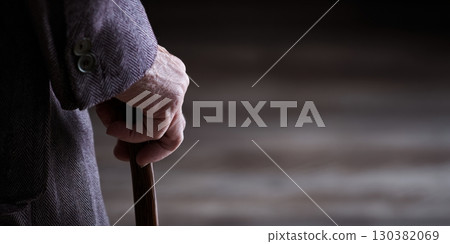 An elderly individual stands quietly, gripping a cane, showing signs of discomfort in a dimly lit space 130382069