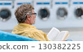 Man with gray hair is reading a book while sitting on a blue couch in a laundromat, surrounded by washing machines and a relaxed atmosphere 130382225