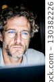 Focused man with curly hair wearing glasses, deeply engaged in work on a laptop, illuminated by soft light in a dark environment 130382226