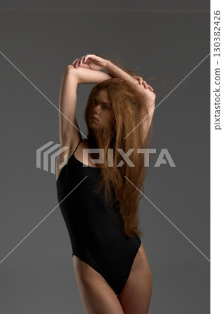 Young woman with long red hair posing in black swimsuit against gray background, expressing elegance and confidence. Young woman with long red hair posing in black swimsuit against gray background, expressing elegance and confidence. 130382426