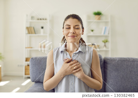 Woman sitting at home with her hands on chest with closed eyes and grateful face expression. Woman sitting at home with her hands on chest with closed eyes and grateful face expression. 130382465