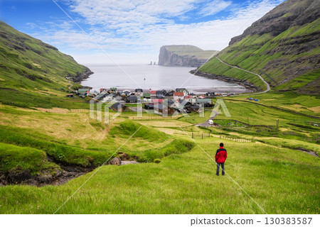 Tourist In Red Jacket Looking At Tjornuvik Village And Ocean, Faroe Islands Tourist In Red Jacket Looking At Tjornuvik Village And Ocean, Faroe Islands 130383587