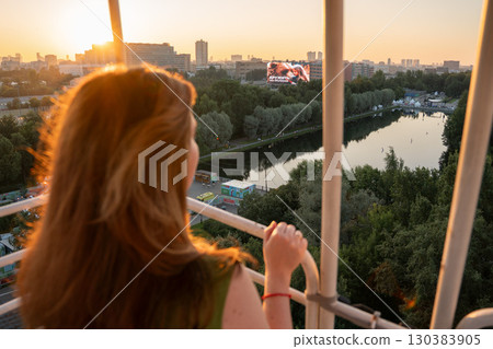 Woman with long hair enjoys sunset view over cityscape and river from a Ferris wheel cabin, warm golden light highlighting the scene 130383905