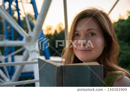 Woman holding an open book looks at the camera while sitting outdoors near a ferris wheel structure at sunset 130383909