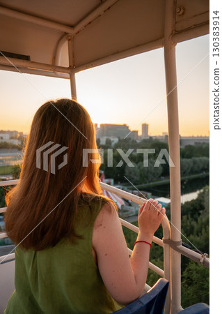 Woman with long hair enjoys sunset view over cityscape and river from a Ferris wheel cabin, warm golden light highlighting the scene 130383914