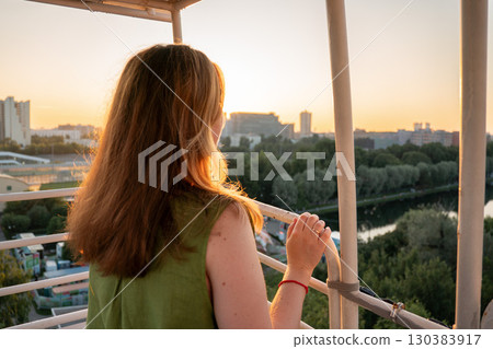 Woman with long hair enjoys sunset view over cityscape and river from a Ferris wheel cabin, warm golden light highlighting the scene 130383917