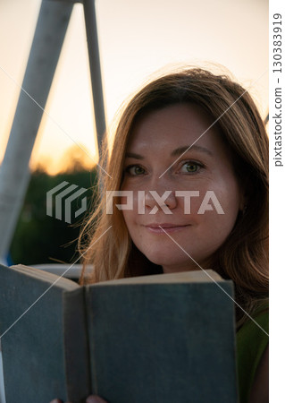 Woman holding an open book looks at the camera while sitting outdoors near a ferris wheel structure at sunset 130383919