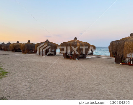 Thatched cabanas line the sandy beach under the pastel evening sky. Coastal leisure, tourism and seaside relaxation with wooden shelters for rest, comfort, and summer vacation ambiance. Thatched cabanas line the sandy beach under the pastel evening sky. Coastal leisure, tourism and seaside relaxation with wooden shelters for rest, comfort, and summer vacation ambiance. 130384295